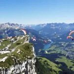 Parapentes colorés, montgolfières, hélicoptère, montagnes alpines, ciel bleu clair, vallée.
