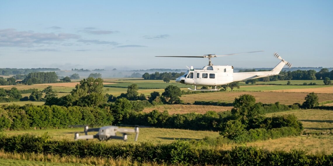 Hélicoptère blanc en vol bas au-dessus du bocage, un petit drone en vol stationnaire au premier plan (illustration)