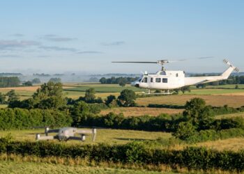 Hélicoptère blanc en vol bas au-dessus du bocage, un petit drone en vol stationnaire au premier plan (illustration)