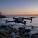 Drone compact en vol au crépuscule au-dessus d’une côte rocheuse, phare en arrière-plan
