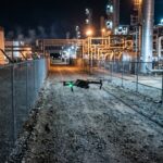 Petit drone volant de nuit le long de la clôture grillagée d’un site industriel éclairé (illustration)