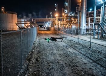 Petit drone volant de nuit le long de la clôture grillagée d’un site industriel éclairé (illustration)