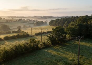 Ligne électrique moyenne tension traversant un bocage au lever du soleil, avec un petit drone qui longe les câbles