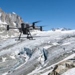 Drone équipé d’une antenne radar survolant un glacier alpin crevassé, deux alpinistes en veste orange au bord de la glace (illustration)