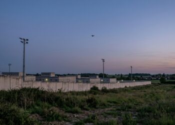 Drone prison : mur d’enceinte d’un établissement pénitentiaire au crépuscule, silhouette lointaine d’un petit drone dans le ciel (illustration)