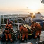 Techniciens en combinaison orange préparant des sacs de matériel sur le pont d’un navire de service, éoliennes en mer à l’aube