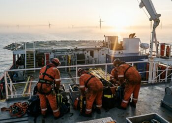 Techniciens en combinaison orange préparant des sacs de matériel sur le pont d’un navire de service, éoliennes en mer à l’aube