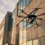 Drone de nettoyage hexacoptère pulvérisant un revêtement sur la façade en béton d’un immeuble de bureaux au coucher du soleil